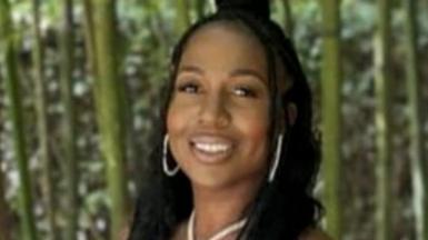 A young woman wearing gold jewellery smiles. She is standing in front of a forest.