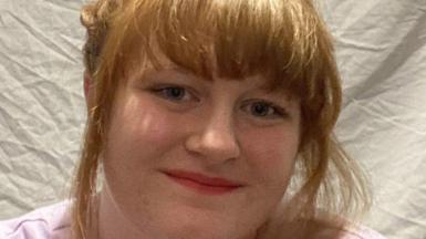 Maisy Moazzenkivi with her short red hair tied up smiling at the camera and wearing a pink t-shirt