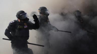 Officers wear riot gear and hold batons as they walk through smoke.