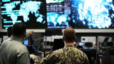 Two us cyber command officers in front of giant screens with data and information on them