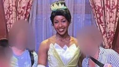 A woman in a blue-striped shirt, with her face blurred, stands next to Disney worker dressed as Princess Tiana in a yellow gown. The woman's daughter, face also blurred, stands on the other side of the princess. The daughter is wearing Mickey Mouse ears and a pink t-shirt