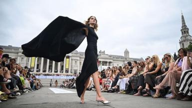 A woman dressed in a black dress waves the back of the dress behind her on a catwalk in Trafalgar Square, with crowds either side of her watching on.