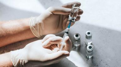 A gloved medical professionals' hand uses a syringe to extract liquid from some vials on a table.