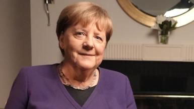 Angela Merkel looks directly into the camera in a portrait image in front of a fireplace