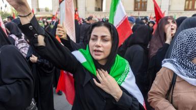 A young Iranian woman in Tehran with an Iranian flag wrapped around her shoulders raises her arm in a crowd of other peple shouting and waving flags