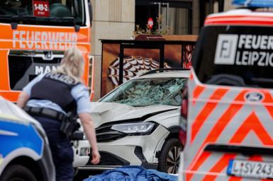 A police officer standing in front of a white car with a smashed windscreen surrounded by fire trucks