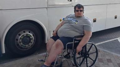 Charley Jonstone-Brent who uses a mobility aid, sitting in a wheelchair in front of a coach. It's a bright day and he is wearing sunglasses, shorts and a t-shirt.