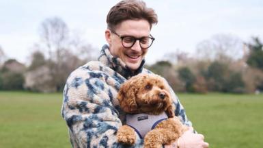 Dr Alex George wearing a blue and white fleece jacket, holding his brown cavapoo rescue dog Rolo