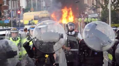 Police officers with riot shields and batons raised, while in the background a police van in burning