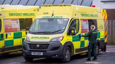 A female paramedic opening the door to an ambulance. Another ambulance is parked alongside.