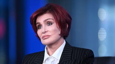 Sharon Osbourne, with trademark red bob, stares into the middle distance while wearing a pinstripe suit.