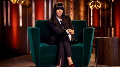 Claudia Winkleman sits on a green velvet sofa wearing a burgundy blazer and trousers