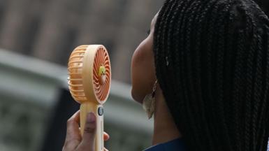 A young woman holds a battery-operated fan close to her face. 
