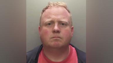 Robert Bush with short, light brown hair is shown in a straight‑on mugshot photograph. He has a neutral expression and is wearing a dark jacket over a red shirt. The background is plain and grey.