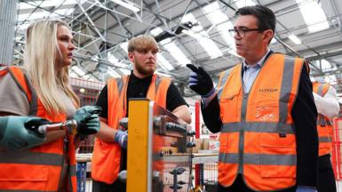 Andy Burhnam wears a hi-viz jacket while talking to two young workers at a factory, also wearing hi-viz jackets, one female with long blonde hair and one man with blonde hair and a beard.