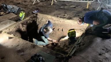Archaeologists at a site are digging in a rectangular hole in the ground surrounded by metal fences. They are wearing head torches