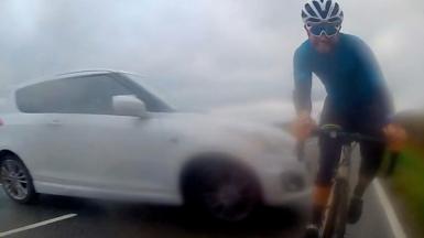 A white car slides sideways across a road, narrowly missing a cyclist who is facing the camera.