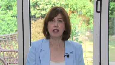 Lucy Powell wearing a light blue jacket and a white top. She is sitting in front of windows that look out on to a garden