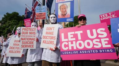 Campaigners from both sides of the debate protest outside Parliament in June 2025, while MPs debate the bill in the House of Commons. One woman holds a pink placard reading: "Let us choose. Legalise assisted dying". Three people wearing white masks and medical gowns holds placards reading: "Don't make doctors killers" and "Protect our NHS from becoming the national suicide service". 