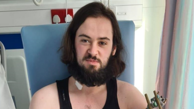 A young man, with long dark brown hair and a brown beard and moustache , sits next to a hospital bed. He has a bandage on his neck.