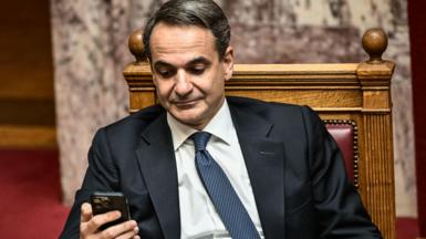 Greek Prime Minister Kyriakos Mitsotakisis wearing a suit and sitting down, looking at a cellphone he holds in his right hand