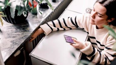 A high angle view of young woman wearing a white and black striped jumper who is checking the temperature of a radiator next to a window. She is looking at a heating app on her smart phone.