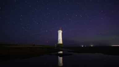 Dark blue night sky showing green and purple colours of the Aurora Borealis visible as a diagonal line of Perseid meteor passed over a white lighthouse 