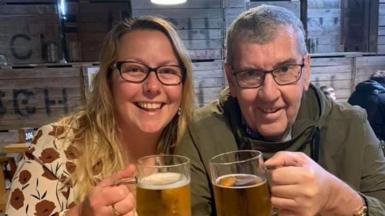 Maureen (left) and Tony Dynes in a pub, smiling at the camera and holding up pints of beer. Maureen has long blonde hair and is wearing a patterned cream top with red flowers. Tony has short grey hair and is wearing a green jacket. Both are wearing glasses.