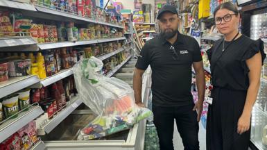 Nazir Ali and Samantha Veira stand in the aisle of a grocery store. Nazir is wearing a black polo shirt and dark pants, while Samantha is wearing a black dress. The aisle is filled with various food products on shelves, including canned goods, sauces, and snacks. A plastic bag filled with confiscated items sits atop a freezer in front of them.