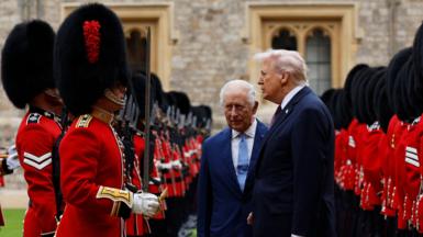 President Donald Trump and King Charles inspect a Guard of Honour together. The pair, both wearing dark suits, are stood between two rows of Beefeaters, dressed in red and with tall black headwear. 