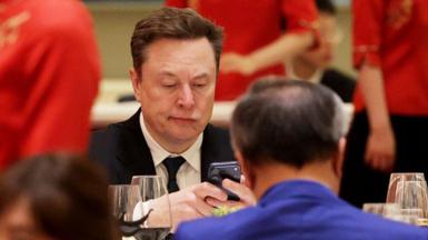 A man in a suit and tie stares at his phone while sitting down opposite a man in a blue jacket in Beijing