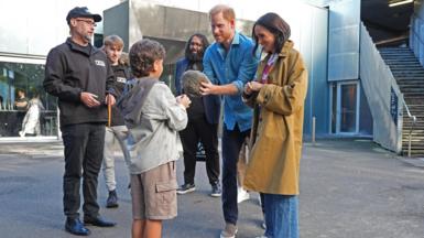 A small boy faces the Duke and Duchess of Sussex, as Prince Harry holds a fur ball and others surround them watching on.