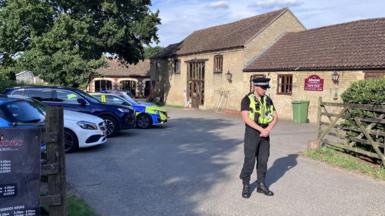 A uniformed police office stands in front of a bricked barn has a car park outside the front on the left. There is a fence on the right which opens into a court yard and there are signs on the door. There is a police car parked in the car park.
