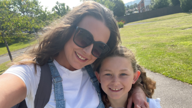 A selfie image of a woman stood in a park in the sunshine with her arm around a young girl. They are both wearing white t shirts and blue denim dungarees. They both have wavy brown hair and the woman is also wearing sunglasses and a grey backpack over one shoulder. behind them is grass and trees. They are both smiling and looking at the camera.