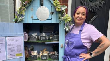 Dani wearing a purple apron and purple t-shirt standing next to the cake shed. The door is open and there are three shelves of cakes. The shed is blue and has a floral garland on the top