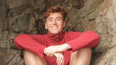 Tomos Sleep who has short light brown hair wearing a red jumper and shorts sat down. 