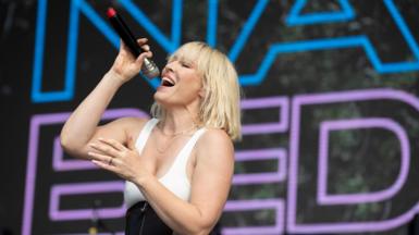 Natasha Bedingfield performs at All Together Now Festival 2024. Natasha is a white woman in her 40s with a bleached blonde bob. She wears a white strappy top and high-rise black trousers. She holds a microphone up with her right hand as she sings, her eyes closed and head titled up.  