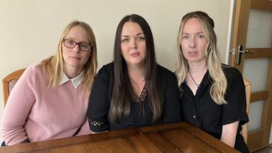 Three women sat at a kitchen table in a row. They are looking at the camera and have serious looks on their faces.