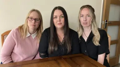 Three women sat at a kitchen table in a row. They are looking at the camera and have serious looks on their faces.