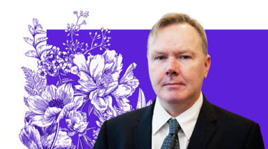 A man in a black suit with a white shirt and blue tie. He has short blond hair and is in front of a purple background with flowers