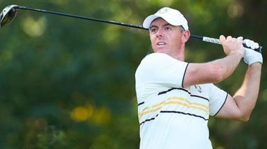 Rory McIlroy watches his ball after hitting a shot with a wood. He is wearing a European Ryder Cup outfit - a baseball cap and a predominantly white polo shirt with black and yellow narrow hoops and trim.