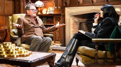 Alan Carr talking to Claudia Winkleman sitting in armchairs in from of a fireplace. There is a pile of gol coins on the table