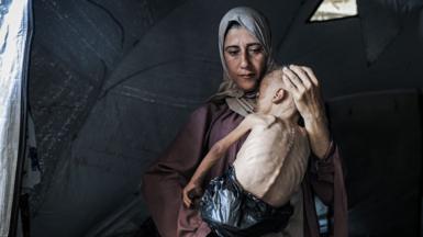 The photograph of a woman holding a famished-looking baby in her arms and looking down at the floor - the baby's face is turned away from the camera and his back is in view, with his spine and ribs visible due to starvation while wearing a black refuse bag as a nappy, in their tent in Gaza City on 21 July.