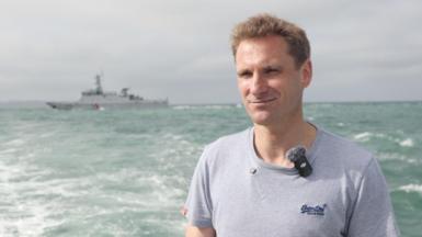 MP Chris Philip, in a grey t-shirt, stands on a boat in the sea. A large, grey ship is visible in the background.