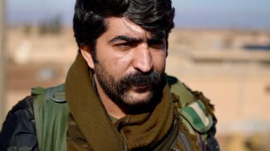 Commander Azad of the Kurdish Forces stares past the camera. He wears a thick black moustache, heavy stubble, and his uniform. 
