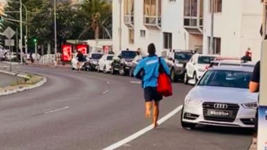 A lifeguard runs down a road barefoot carrying a red defribrillator bag next to parked cars on a road with trees and the sea in the background.