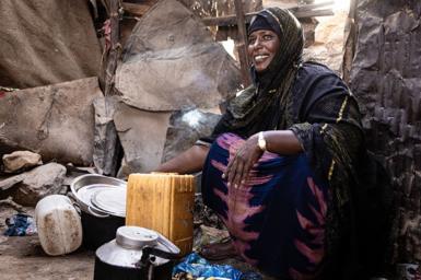 A Somali woman crouching in a makeshift tent, smiles. She is surrounded by pans and water containers,