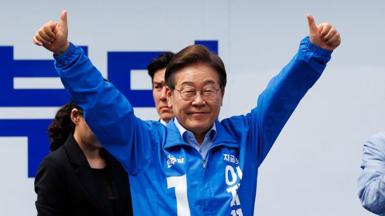 Lee Jae–myung in a blue tracksuit jacket and glasses lifts both hands into the air with thumbs raised
