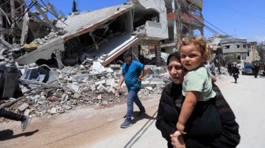 A woman carries a small girl while walking down a street in front of a destroyed building 