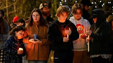 A row of five teenage mourners light candles near the scene. It is night time